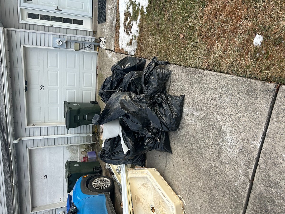 Black garbage bags and items stacked on a residential driveway next to a white garage door